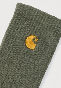 Ελιά πράσινη κάλτσα με ριμπ υφή· διαθέτει ένα κίτρινο κεντητό λογότυπο στροβίλου στο πλάι. Το υλικό φαίνεται μαλακό και ανθεκτικό.