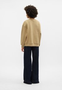 Beige oversized mikina s kleslými rameny, ribovanými manžetami a klasickým kulatým výstřihem, spárovaná s tmavě modrými širokými džíny.