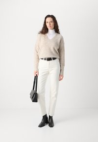 Beige teksturert genser med v-hals, hvit turtleneck under, hvite jeans, svart belte og svarte ankelstøvler, holder en liten svart veske.