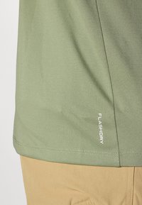 Zbliżenie osoby ubrane w jasnozieloną sportową koszulkę z napisem "FLASHDRY" przy dolnej krawędzi oraz beżowe spodnie.