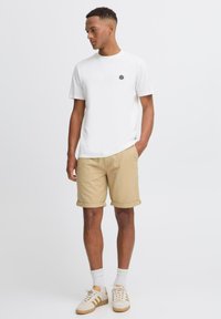 Weißes T-Shirt mit kurzen Ärmeln und schwarzem Logo, kombiniert mit beigen umgeschlagenen Shorts, weißen Sneakers mit beigen Akzenten und weißen Socken.