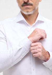 Camicia da uomo bianca a maniche lunghe con colletto classico, polsini con bottoni e tessuto liscio, mostrata con mani che aggiustano i bottoni dei polsini.