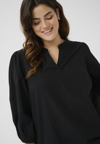 Schwarze Bluse mit V-Ausschnitt, puffärmeligen Ärmeln und vertikalem Streifenmuster. Der Stoff wirkt glatt mit einem leichten Glanz und subtiler Textur.