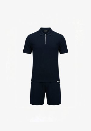 Man staande, draagt een zwart poloshirt met korte mouwen en rits, zwarte shorts en zwarte bandjes sandalen op een effen witte achtergrond.