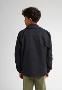 Jachetă neagră cu o croială lejeră, guler cu vârf, mâneci lungi și manșete elastice, purtată cu pantaloni verzi deschis. Fără modele sau accente vizibile.