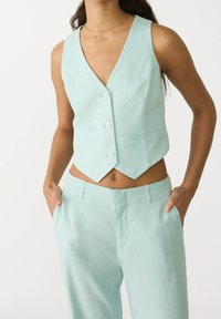 Chaleco sin mangas verde menta con cuello en V, con tres botones y dos bolsillos laterales, combinado con pantalones a juego en verde menta.