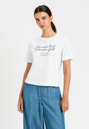 Giovane donna con capelli scuri raccolti in una coda di cavallo, indossa una maglietta bianca con scritta viola e pantaloni di jeans blu larghi, in piedi davanti a uno sfondo semplice.