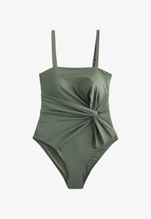 Primo piano di un costume da bagno verde con un nodo intrecciato in vita e una mano che tiene delicatamente il tessuto su uno sfondo di spiaggia.