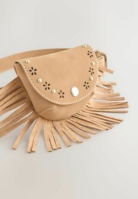 Sac banane en daim camel avec découpes florales, clous dorés et longues franges sur fond clair.