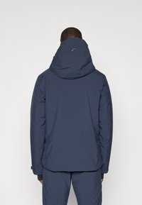 Giacca impermeabile blu navy con cappuccio, dotata di una finitura opaca, polsini regolabili e un dettaglio con il logo sulla schiena.