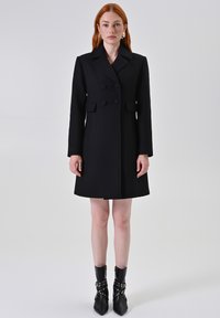Cappotto nero a doppio petto in tessuto texturizzato, con colletto a punta e due tasche frontali. Il modello abbina stivali neri a punta con dettagli in rivetti.