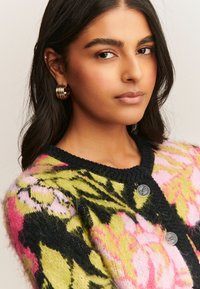 Cardigan en tricot avec des motifs floraux vibrants dans des tons de rose, vert et noir, doté d'une finition texturée et de deux boutons argentés à l'avant.