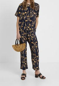 Marinblått tvådelat set med blommönster i gult, bestående av en lös passform överdel och byxor. Modellen håller en bambu halvmåne-väska och bär svarta slides.