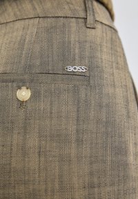 Beige teksturoidut housut, joissa on hillitty ruutukuvio, etutasku, yksittäinen nappikiinnitys ja hopeinen "BOSS"-logo.