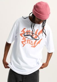 Camiseta blanca de gran tamaño con un diseño gráfico en naranja y gris en el frente. El modelo lleva un gorro de lana rosa y pantalones oscuros.