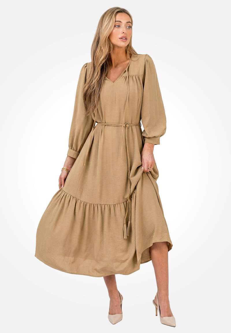 Mujer con un vestido midi de manga larga beige con un cinturón trenzado, sosteniendo la falda y tacones altos puntiagudos beige sobre un fondo blanco.