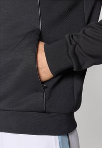 Schwarze Sportjacke mit strukturiertem Oberteil, glattem Unterteil, Reißverschlusstasche und gerippten Bündchen sowie Saum.