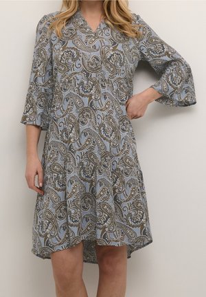 Mujer con un vestido azul claro hasta la rodilla con estampados de cachemira marrones y blancos, con mangas campana 3/4 y cuello en V.