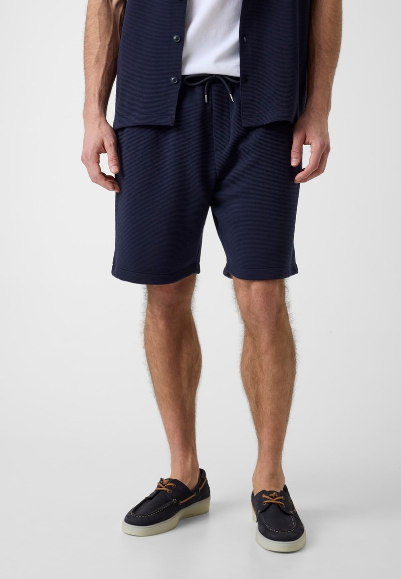 Homme portant un short bleu marine à cordon, une chemise ouverte assortie, un t-shirt blanc et des chaussures bateau noires, debout sur un fond uni.