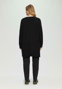 Lange zwarte gebreide trui met een ontspannen pasvorm, voorzien van een ronde halslijn en zijsplitten, gecombineerd met zwarte broek en stevige donkere schoenen.