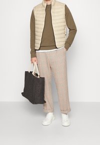 Beige vadderad väst över en olivfärgad sweatshirt, rutiga byxor med röda detaljer, och vita sneakers. Håller i en strukturerad svart tote-väska.
