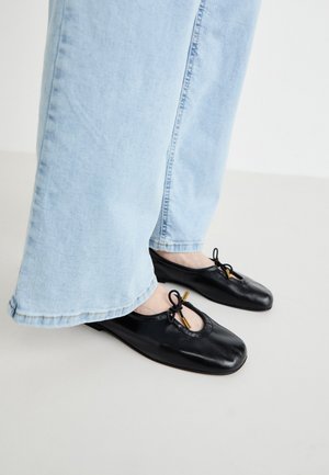 Schwarze Leder-Ballettschuhe mit runder Zehenpartie, die mit einem Ausschnitt und einer Schleifendetail versehen sind, getragen mit hellblauen weit geschnittenen Jeans.