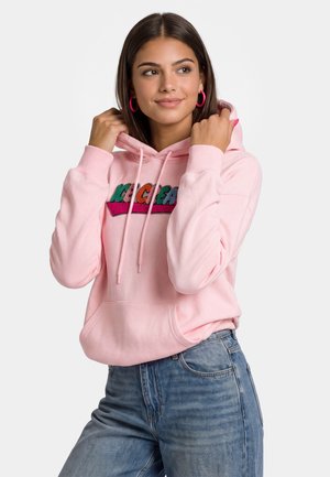 Ung kvinde iført pink hættetrøje med farverig tekst og blå jeans, der holder i hættetrøjens snore, smiler let og kigger til siden.