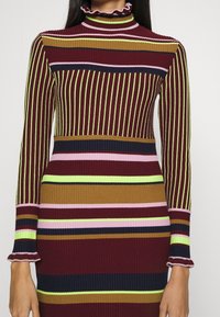 Ribbad långärmad klänning med horisontella ränder i vinröd, gul, marinblå, grön och rosa. Hög volangkrage och figurnära design.