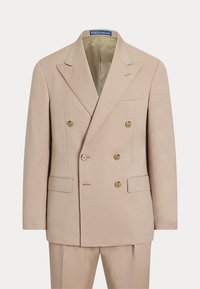 Veste de costume beige croisée avec boutons dorés, revers crantés et poches à rabat, présentée avec un pantalon assorti.