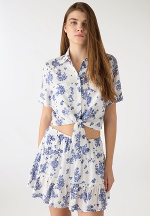 Giovane donna con lunghi capelli castani che indossa un completo composto da una camicia a maniche corte e una gonna con motivi floreali blu, camicia annodata in vita.