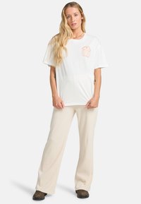 Weißes Kurzarm-T-Shirt mit einer hellorangefarbigen Grafik auf der Brust; kombiniert mit beigen gerippten Hosen und dunklen Schuhen.