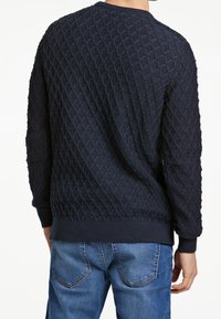 Dunkelblauer gestrickter Pullover mit einem strukturierten Rautenmuster. Mit gerippten Bündchen und Saum ausgestattet. Getragen mit hellblauen Jeans.
