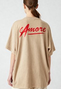 Blaženo oversized T-maica iz khaki barve, izdelana iz mehkega materiala, z velikim rdečim grafičnim napisom "Amore" na hrbtni strani, ki ima gladko teksturo in sproščen kroj.