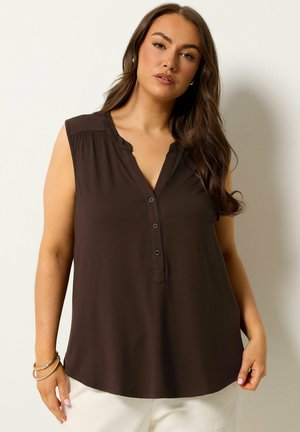 Vrouw met lang bruin haar, die een mouwloze donkerbruine blouse met knopen en een witte broek draagt, staand tegen een effen lichte achtergrond.