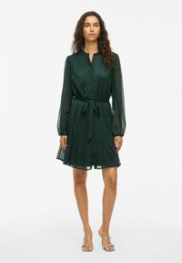 Rochie verde cu guler, mâneci lungi din material transparent, cordon în talie și tiv volanat. Fabricată dintr-un material ușor, prezintă o croială lejeră.