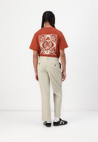 Camiseta de color óxido con diseño geométrico floral blanco en la parte posterior, combinada con pantalones beige claros y zapatillas negras. Textura suave, ajuste casual.