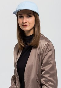 Világoskék sapka ívelt nappal, barna selyem bomberdzseki viselve egy fekete felsőn. Egyenes, közepes hosszú haj látható.