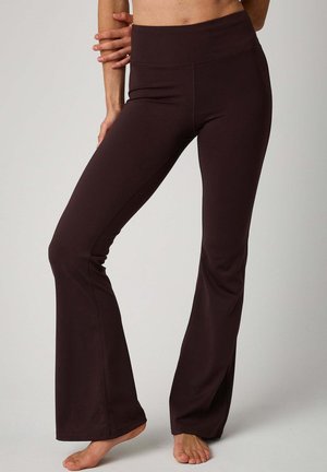 Woman wearing dark brown high-waisted flare yoga pants, standing barefoot with one leg slightly bent and hand resting on her waist.