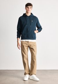 Felpa blu navy con tasca frontale e coulisse, abbinata a pantaloni beige e sneakers bianche, su uno sfondo chiaro.