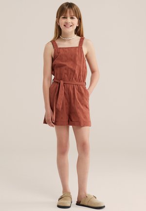 Tüdruk naeratab, seljas roostetooni varrukateta vöödiline kombinesoon lühikeste pükstega ning beežid libisevad jalatsid, seisab ühevärvilise tausta ees.