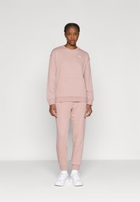 Απαλό ροζ φούτερ με μπροστινή τσέπη και λευκό λογότυπο, σετ με αντίστοιχο παντελόνι φόρμας. Το ύφασμα φαίνεται απαλό και λείο. Λευκά αθλητικά παπούτσια ολοκληρώνουν το ντύσιμο.