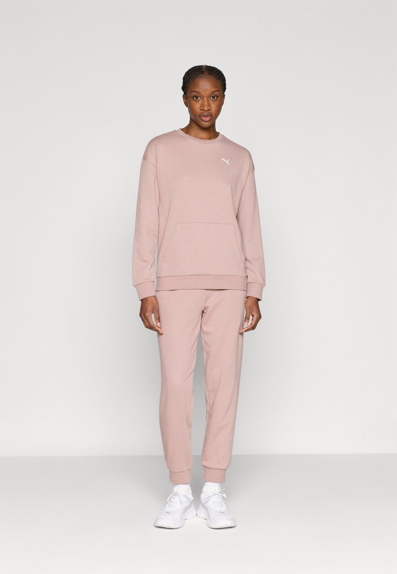 Απαλό ροζ φούτερ με μπροστινή τσέπη και λευκό λογότυπο, σετ με αντίστοιχο παντελόνι φόρμας. Το ύφασμα φαίνεται απαλό και λείο. Λευκά αθλητικά παπούτσια ολοκληρώνουν το ντύσιμο.
