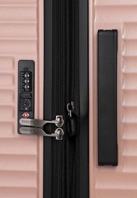 Valigia rigida con cerniera dotata di doppi cursori in metallo, lucchetto a combinazione TSA integrato e maniglia laterale nera su superficie rosa chiaro testurizzata.