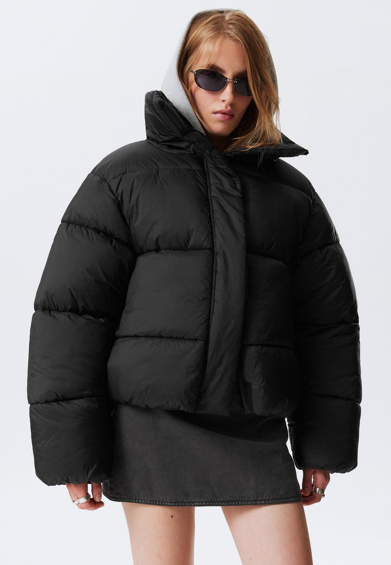 Crni puffer kaput s visokim ovratnikom, oversized oblikom i vidljivim šavovima. Nosite ga preko sive dukserice i uparite s tamnom suknjom.