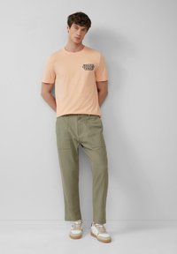 Licht perzikkleurig katoenen t-shirt met zwarte bedrukte tekst, gecombineerd met olijfgroene taps toelopende broek en witte sneakers met beige accenten.