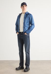 Blauer Nylon-Windbreaker mit Kapuze, beiges T-Shirt, dunkelblaue Jeans und schwarze Slipper. Glatte Textur mit Reißverschluss.