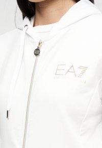 Κοντινή εικόνα ατόμου που φοράει λευκό φούτερ με φερμουάρ EA7 Emporio Armani με λογότυπο σε μεταλλικά τρουκς στο στήθος και ασημένια ταινία φερμουάρ.