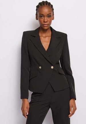 Donna con capelli intrecciati che indossa un blazer doppiopetto nero aderente e pantaloni su misura abbinati, su uno sfondo bianco semplice.