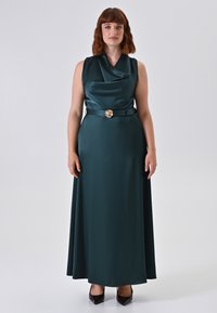 Donna che indossa un abito senza maniche in raso verde scuro con scollatura a cascata e un cinturino con fiore dorato, in piedi davanti a uno sfondo chiaro e uniforme.