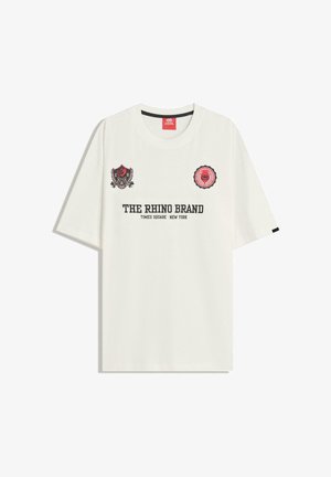 Bela bomba majica iz bombaža z natisnjenim črnim in rdečim besedilom, dvema emblemoma na zgornjem sprednjem delu in krožno izrezano obliko. Kratke rokave.